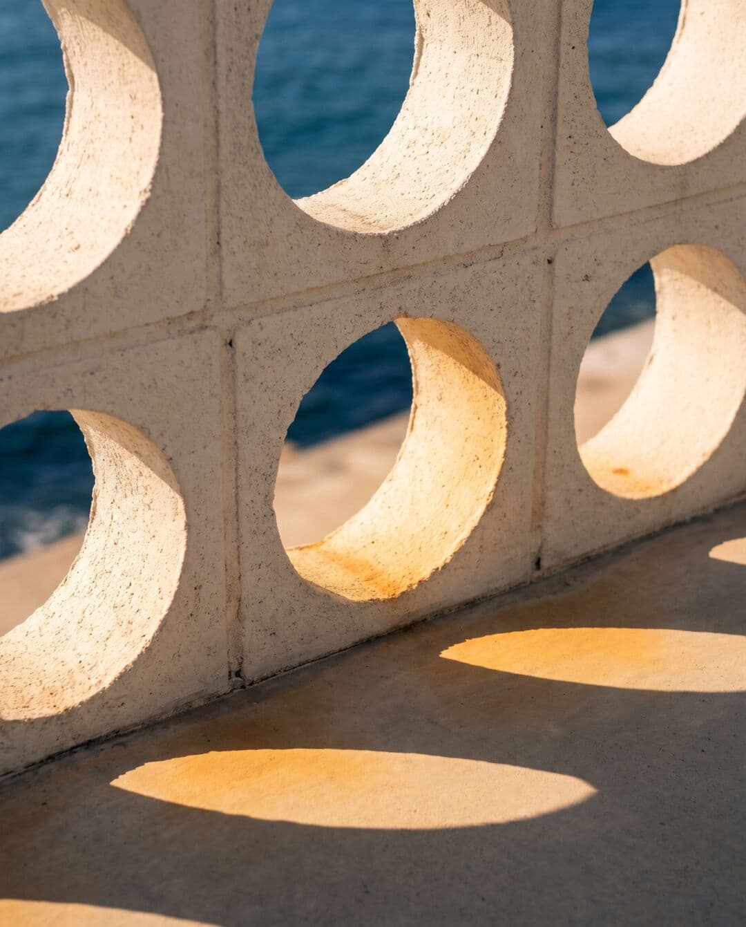 Primo piano di una claustra in cemento con aperture circolari attraversata dal sole del tardo pomeriggio, sullo sfondo il mare